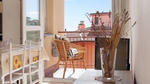 Un balcón con una silla de mimbre, una mesa con sillas y un florero con plantas secas.