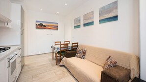 A modern kitchen with white cabinets, a dining area with a table and chairs, a beige sofa with patterned cushions, and three wall-mounted paintings depicting landscapes. - Vacation Apartment Trilocale Recagno nuovo near the Beach with A/C (Varazze)