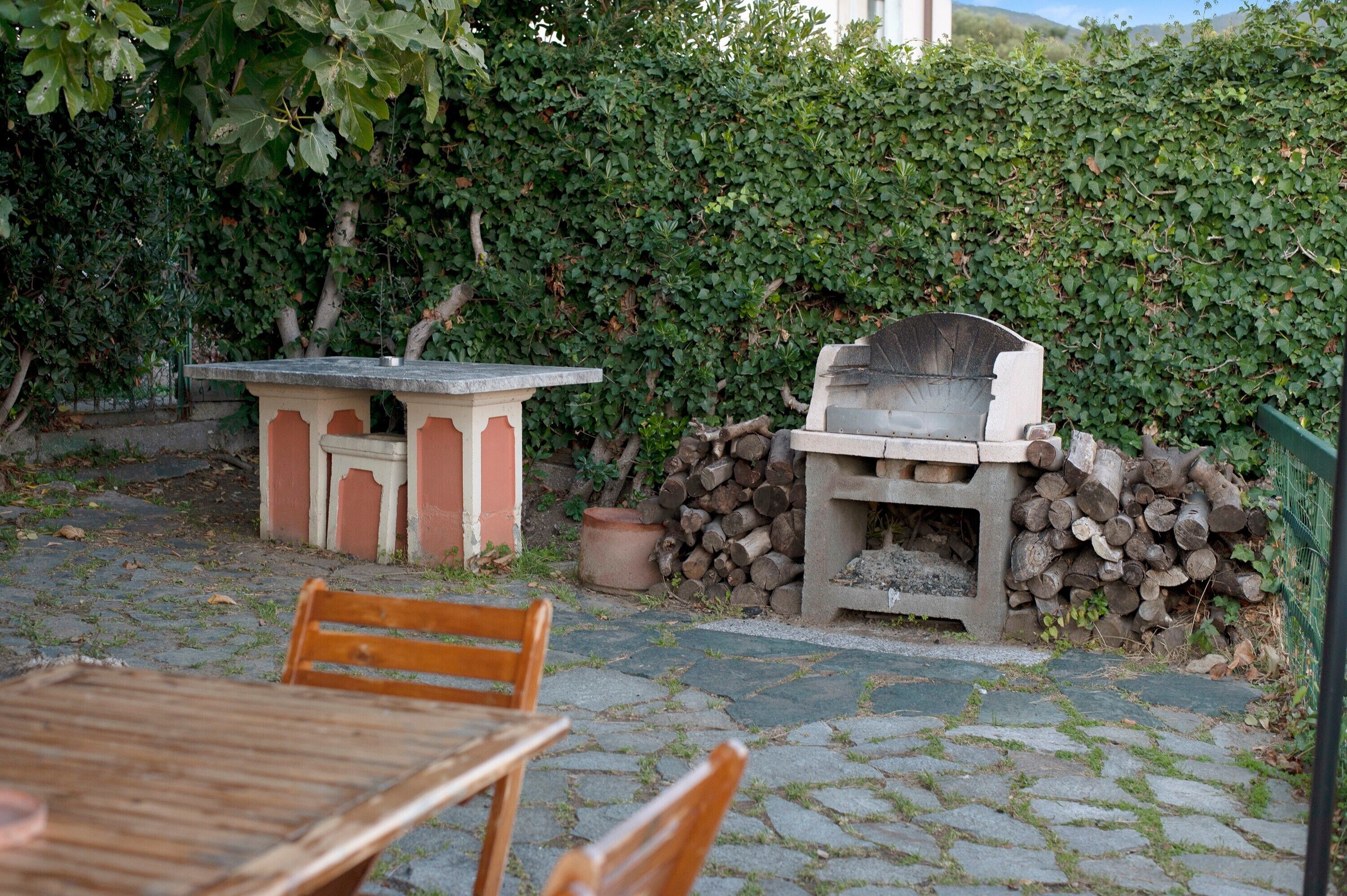 Eine Terrasse im Freien mit einem Steintisch, Holzstühlen, einem Grill und einem Holzstoß.