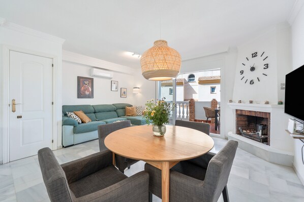 Un salon avec une table ronde en bois, des canapés gris, un luminaire suspendu, une horloge murale et une cheminée.