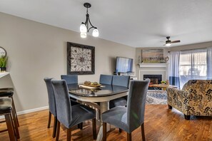 Un coin repas avec une table ronde, six chaises et un lustre. On aperçoit également un téléviseur, une cheminée et un canapé au fond.