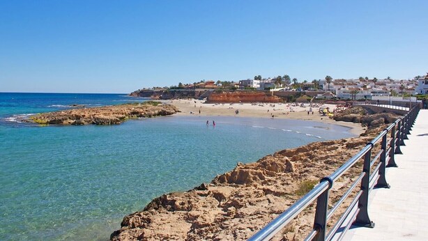 Escena costera de una playa con agua cristalina, un litoral rocoso y un paseo marítimo con barandilla.