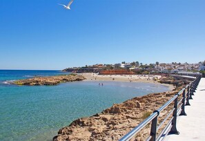 A coastal beach scene with clear water, a rocky shoreline, and a promenade with a railing.