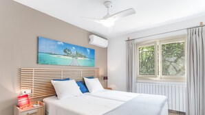 Un dormitorio con cama, mesita de noche, ventilador de techo, ventana con cortinas y un cuadro de una playa.