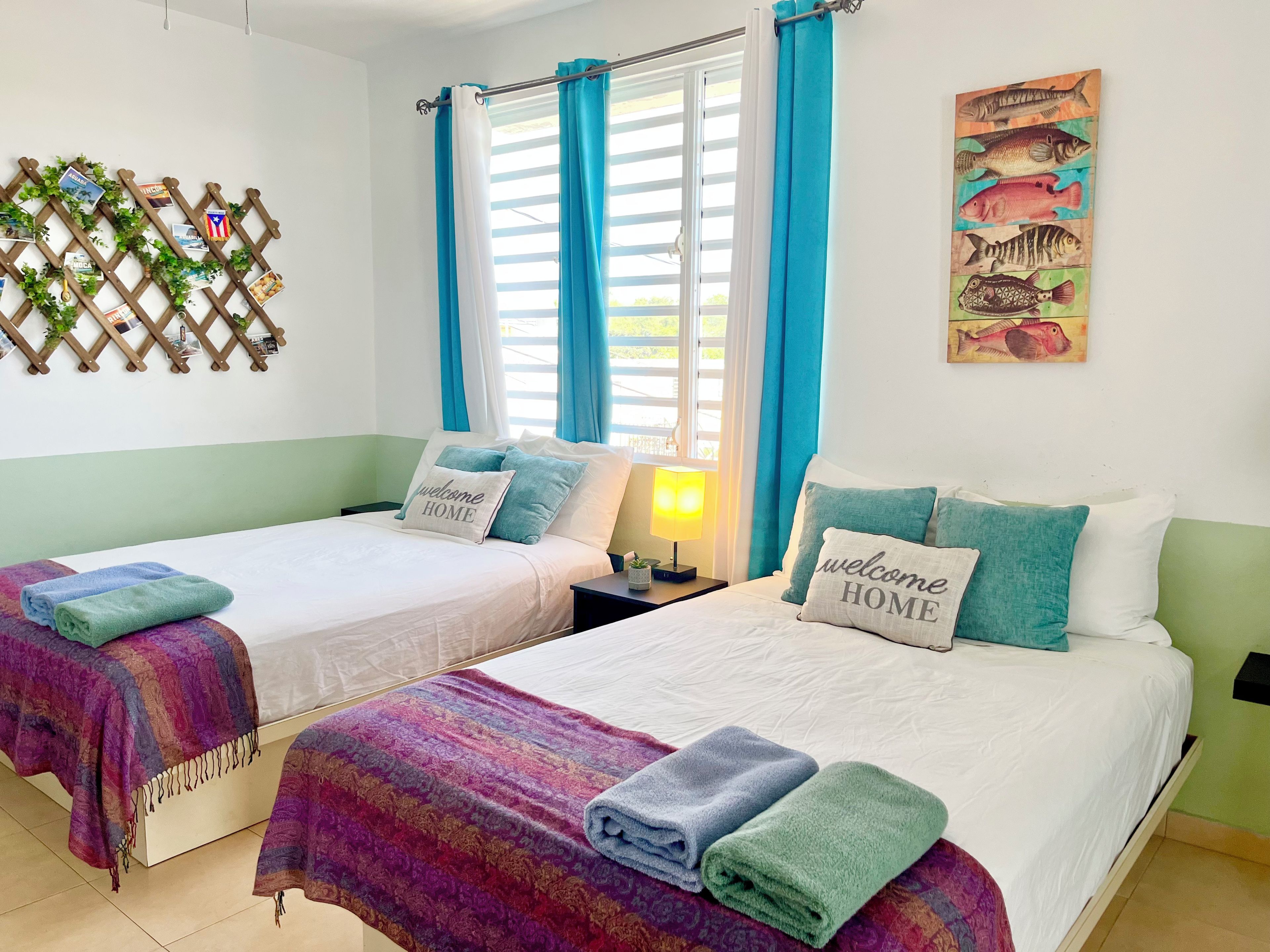 Una habitación de hotel con dos camas, un ventanal con cortinas, una planta en la pared y una obra de arte con motivos marinos.