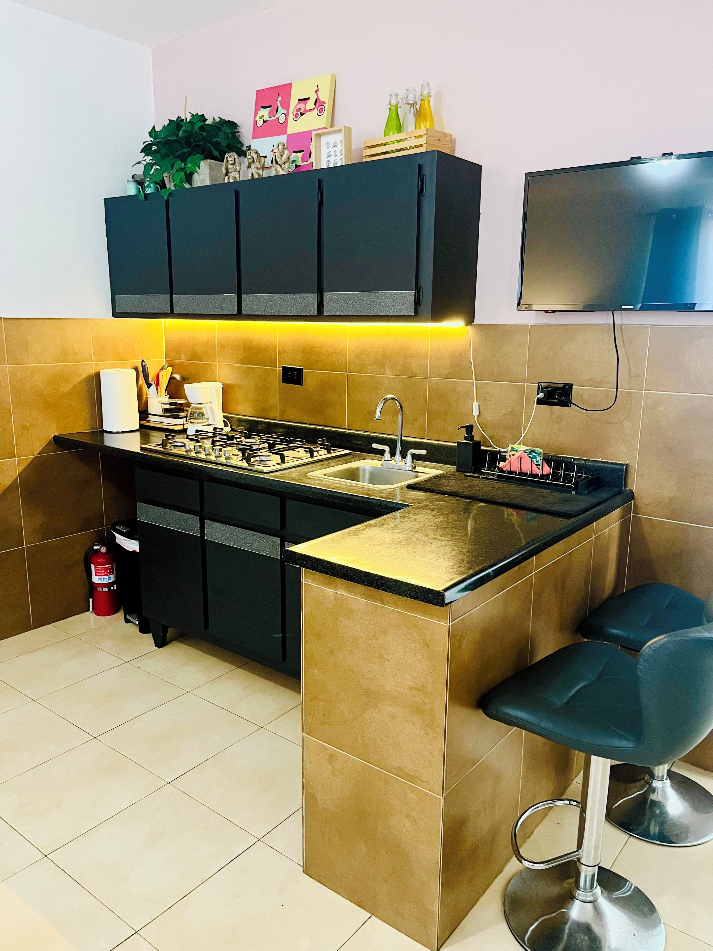 Cocina con isla central, armarios negros, salpicadero azulejado, estufa a gas, un lavamanos y dos taburetes de bar.