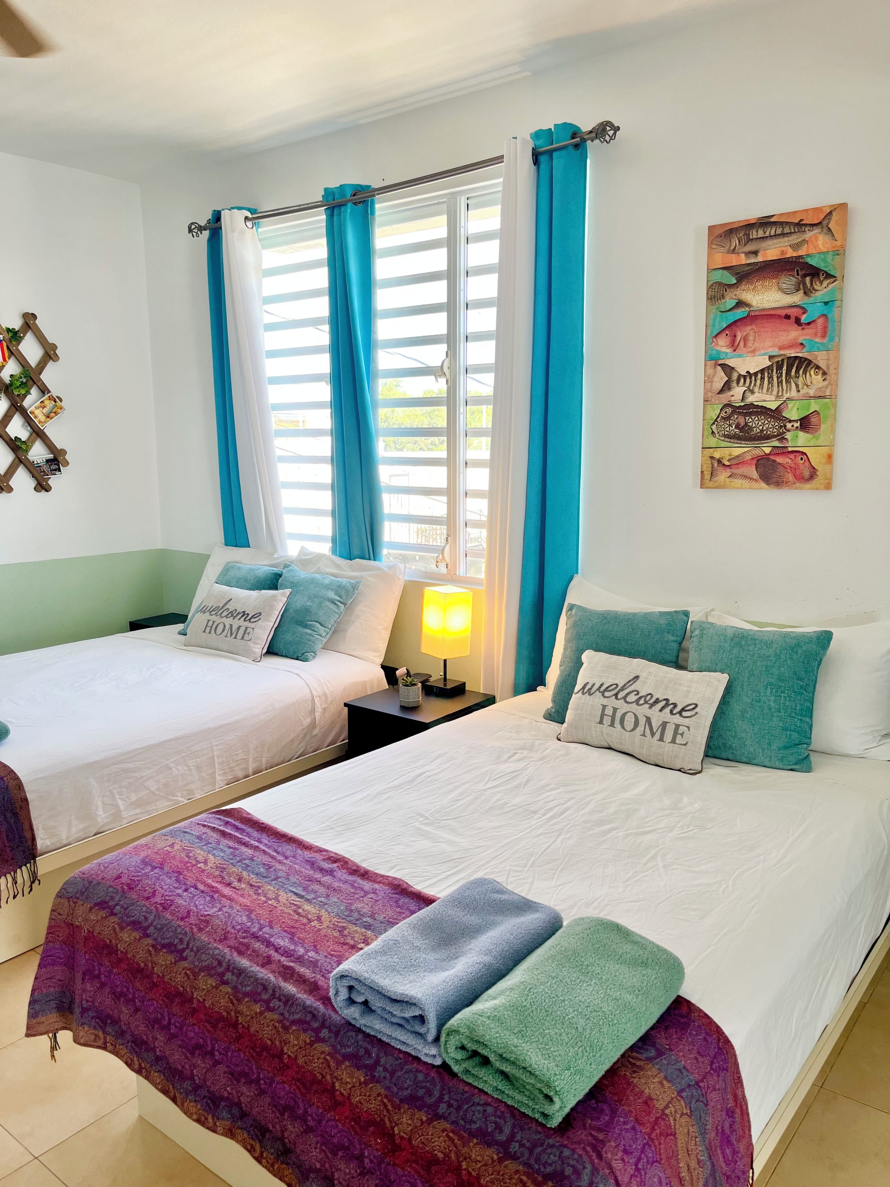 Habitación de hotel con dos camas, una ventana con cortinas, una obra de arte en la pared y una mesita de noche con lámpara.