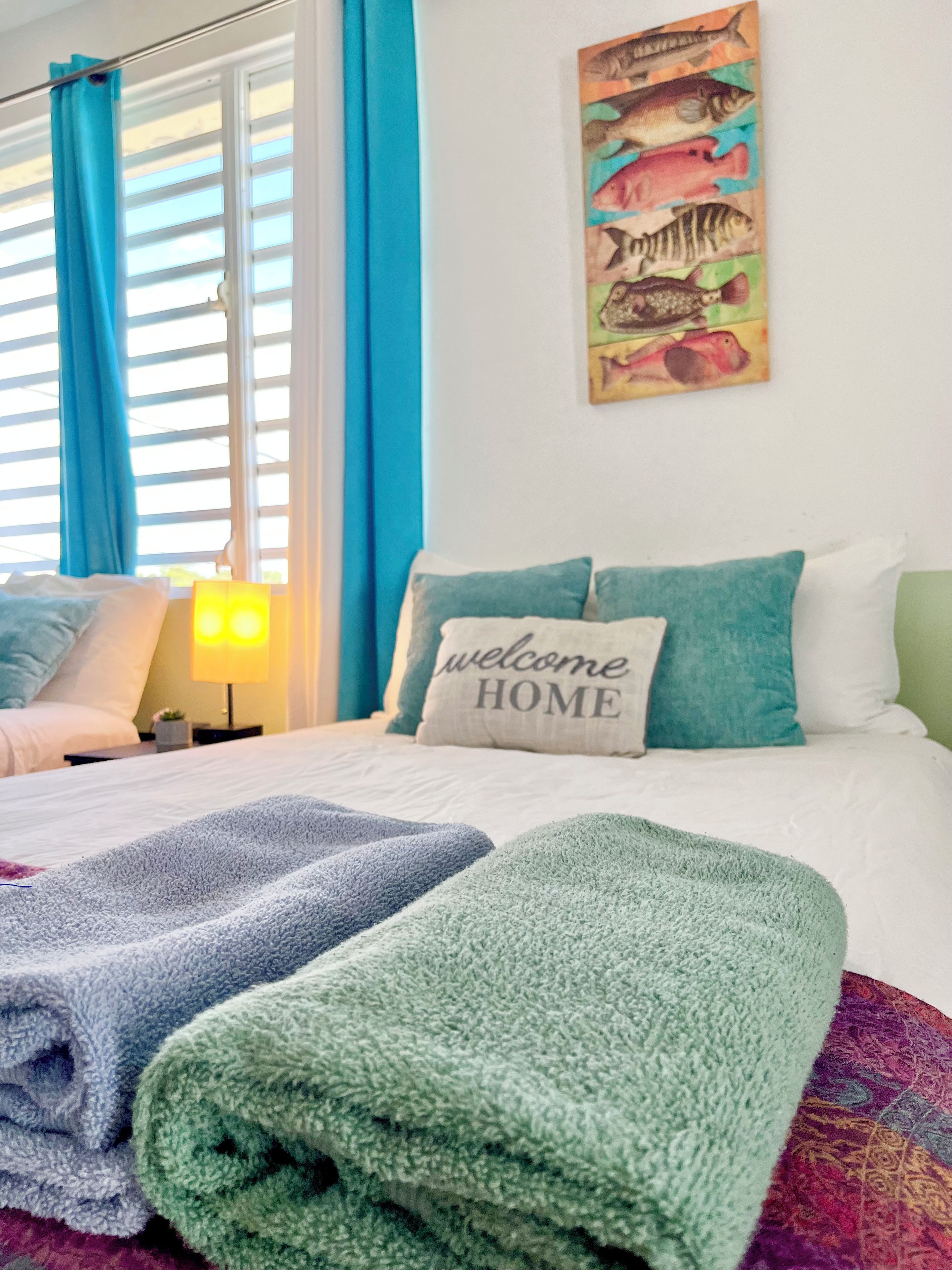 Una habitación de hotel con cama, almohada con el texto 'welcome HOME', un cuadro de peces y una ventana con cortinas.