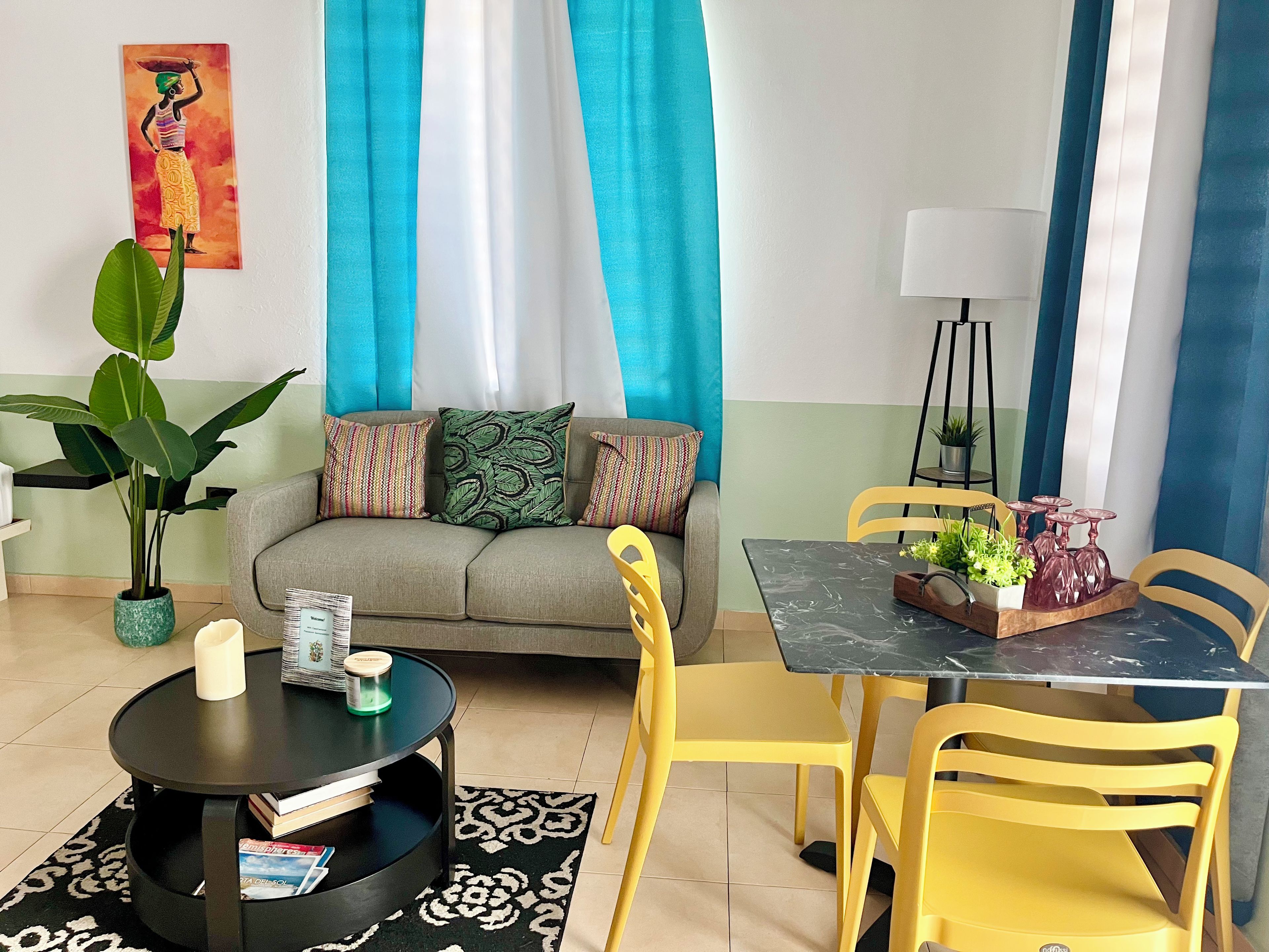 A living room with a sofa, a round coffee table, yellow chairs, a dining table with chairs, a potted plant, and a painting on the wall.