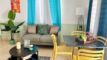 A living room with a sofa, a round coffee table, yellow chairs, a dining table with chairs, a potted plant, and a painting on the wall.