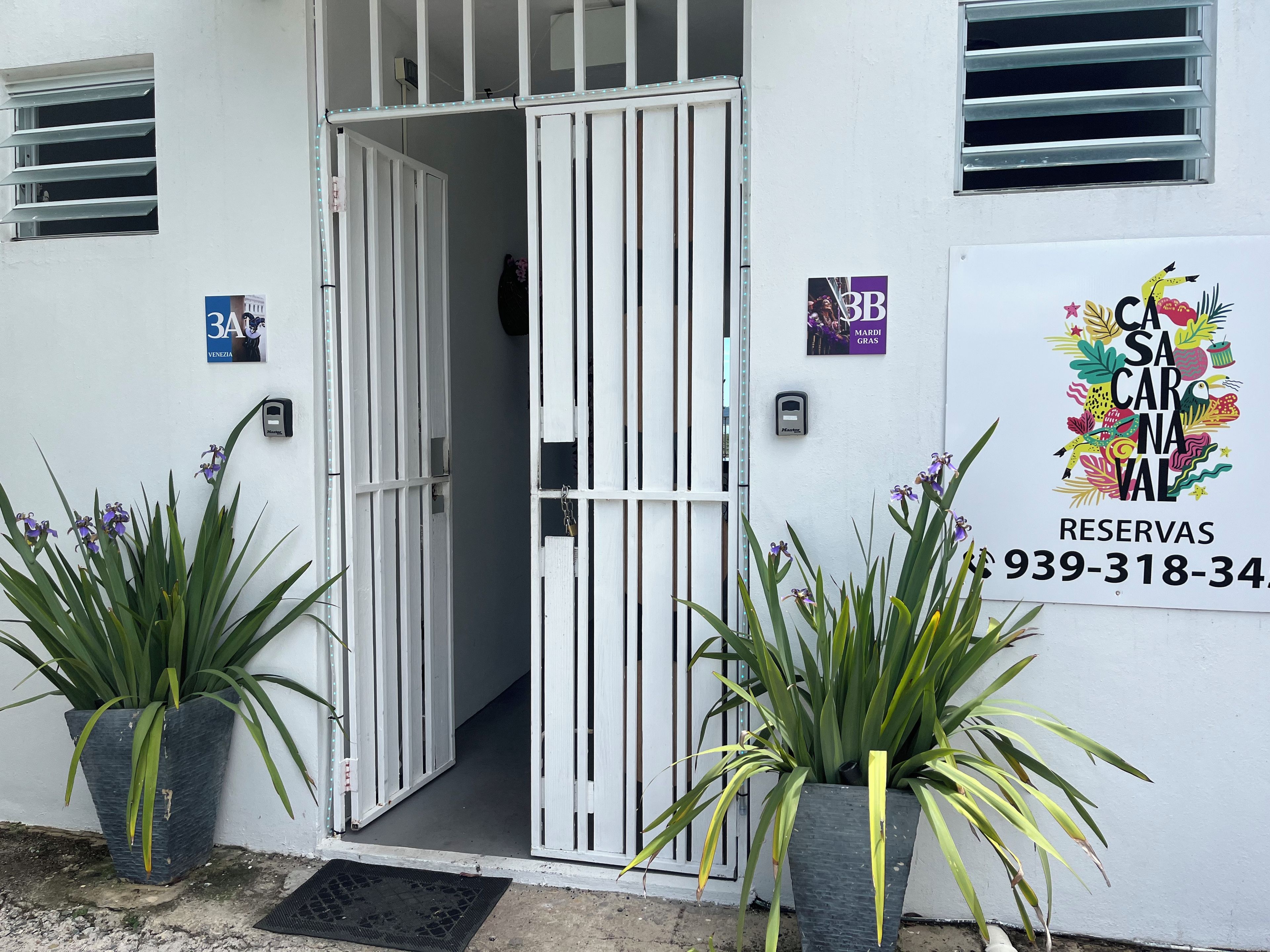 Entrada de hotel con una reja blanca, plantas en macetas y un letrero con un número de teléfono.