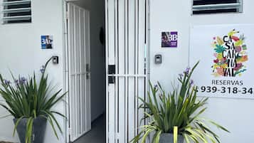 Entrada de hotel con una reja blanca, plantas en macetas y un letrero con un número de teléfono.