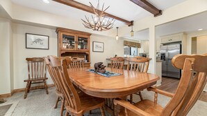Uma sala de jantar com mesa e cadeiras de madeira, um candelabro e uma cozinha ao fundo.
