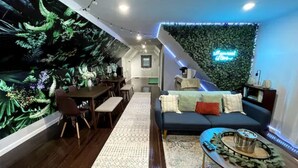 Ein modernes Wohnzimmer mit dunklem Holzboden, einem gemusterten Teppich, einem dunklen Sofa mit Kissen, einem Couchtisch mit einer Dekoration und einer Wand mit einem grünen Blattmuster.