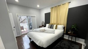 Ein Schlafzimmer mit einem großen Bett, einem dunklen Teppich, einem kleinen Tisch mit einer Pflanze und einer Schiebetür zu einem Außenbereich.