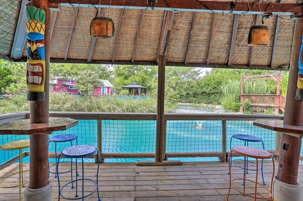 A wooden deck with a pool, blue stools, and a thatched roof.