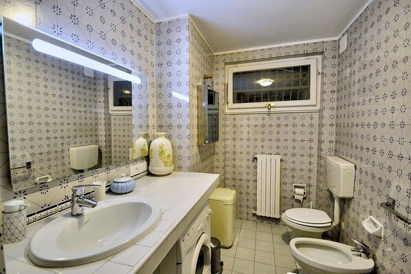Un baño con un lavamanos grande, inodoro y bidé. Presenta papel tapiz estampado y una ventana sobre el inodoro.