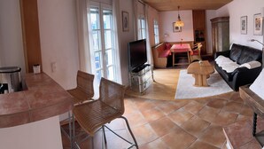 A kitchen with wooden cabinets, a microwave, and a sink. There is a window with curtains, a dining area with a table and chairs, and a living room with a sofa and a TV. - Wohnung Nr. 5 Sonnenrose 56 qm mit einem Schlafzimmer, zwei Bäder und Südbalkon (Bad Reichenhall)