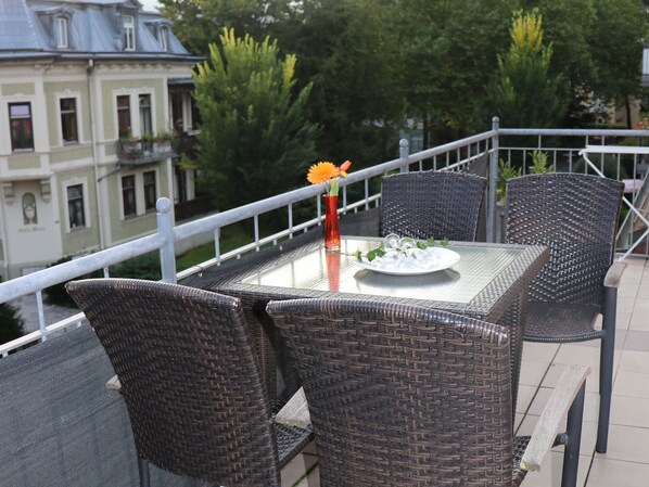 A balcony with wicker chairs and a table, a vase with flowers, and a plate of food.