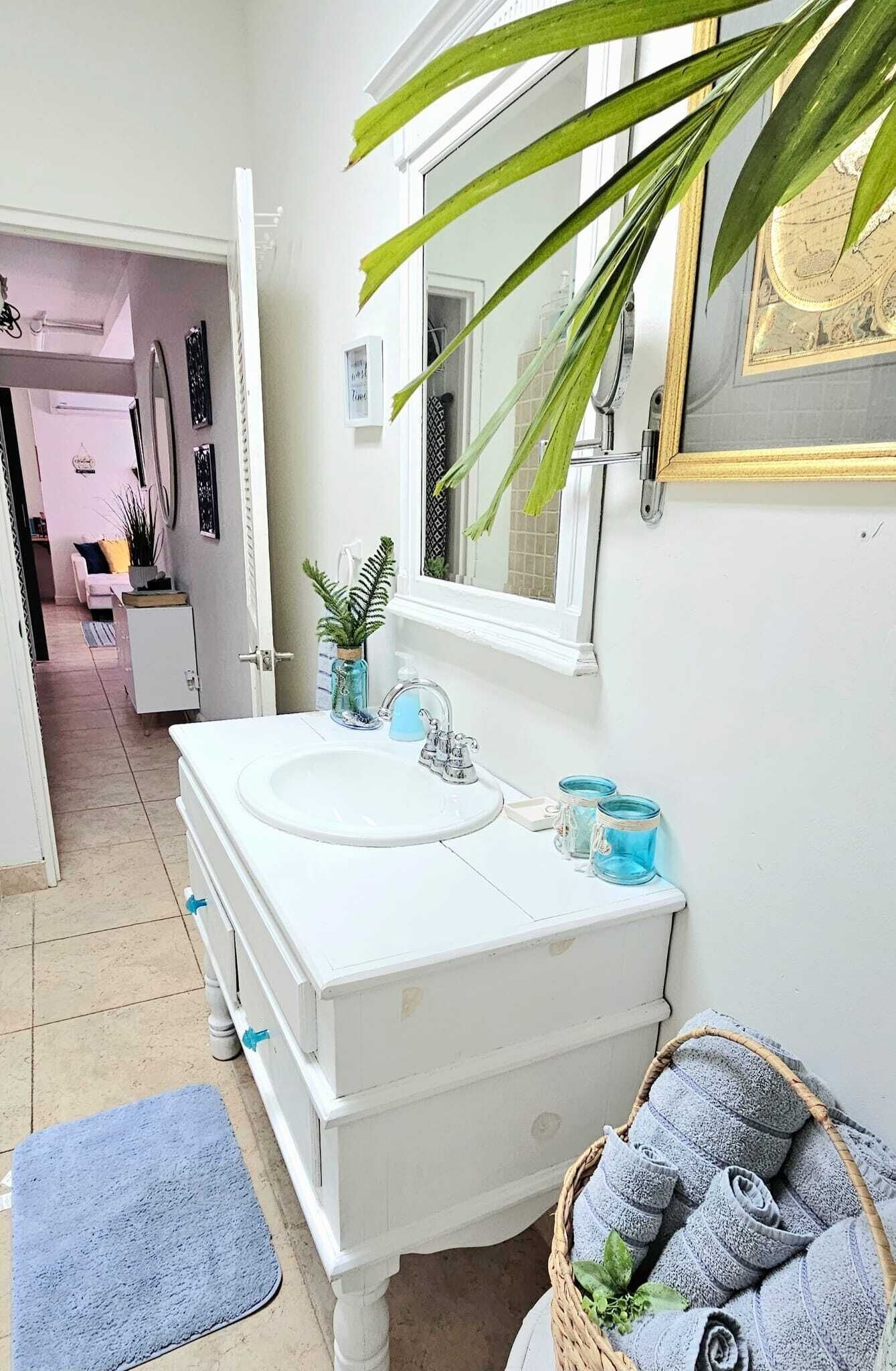 Une salle de bain avec un meuble-lavabo blanc, un évier, un miroir et une fenêtre. On y trouve une plante sur le meuble-lavabo et un panier rempli de serviettes sur le sol.
