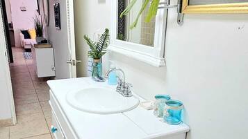 Een badkamer met een wit wastafelmeubel, een wastafel, een spiegel en een raam. Op het meubel staat een plant en op de vloer een mand met handdoeken.
