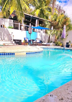Une piscine à l’eau d’un bleu limpide, entourée de chaises longues et de parasols, avec des palmiers et un bâtiment en arrière-plan.