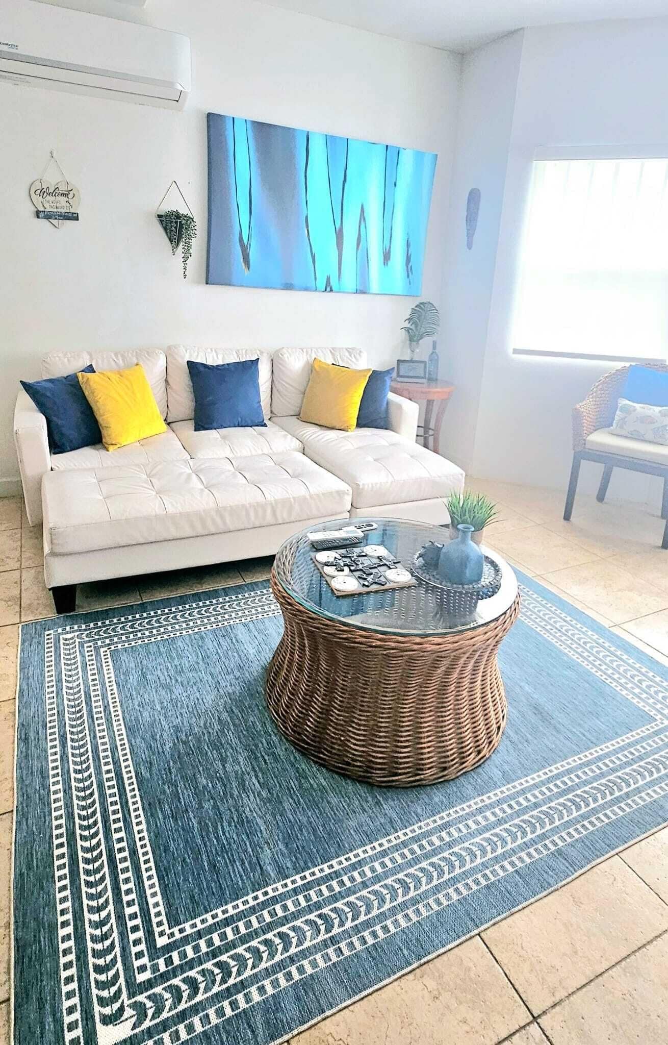 Una sala de estar con un sofá blanco, cojines azules y amarillos, una mesa de centro de cristal con adornos, y una alfombra estampada azul.