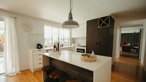 A modern kitchen with a large island, bar stools, a hanging light fixture, and a view of the living area. - Gorgeous 5 bedroom beach house. Dog/Kid Friendly. (Marcoola)
