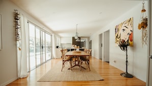 A spacious dining area with a wooden table and chairs, large windows, and a modern kitchen in the background. - Gorgeous 5 bedroom beach house. Dog/Kid Friendly. (Marcoola)