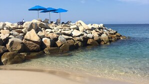Una spiaggia rocciosa con acque limpide, una riva sabbiosa e ombrelloni blu.