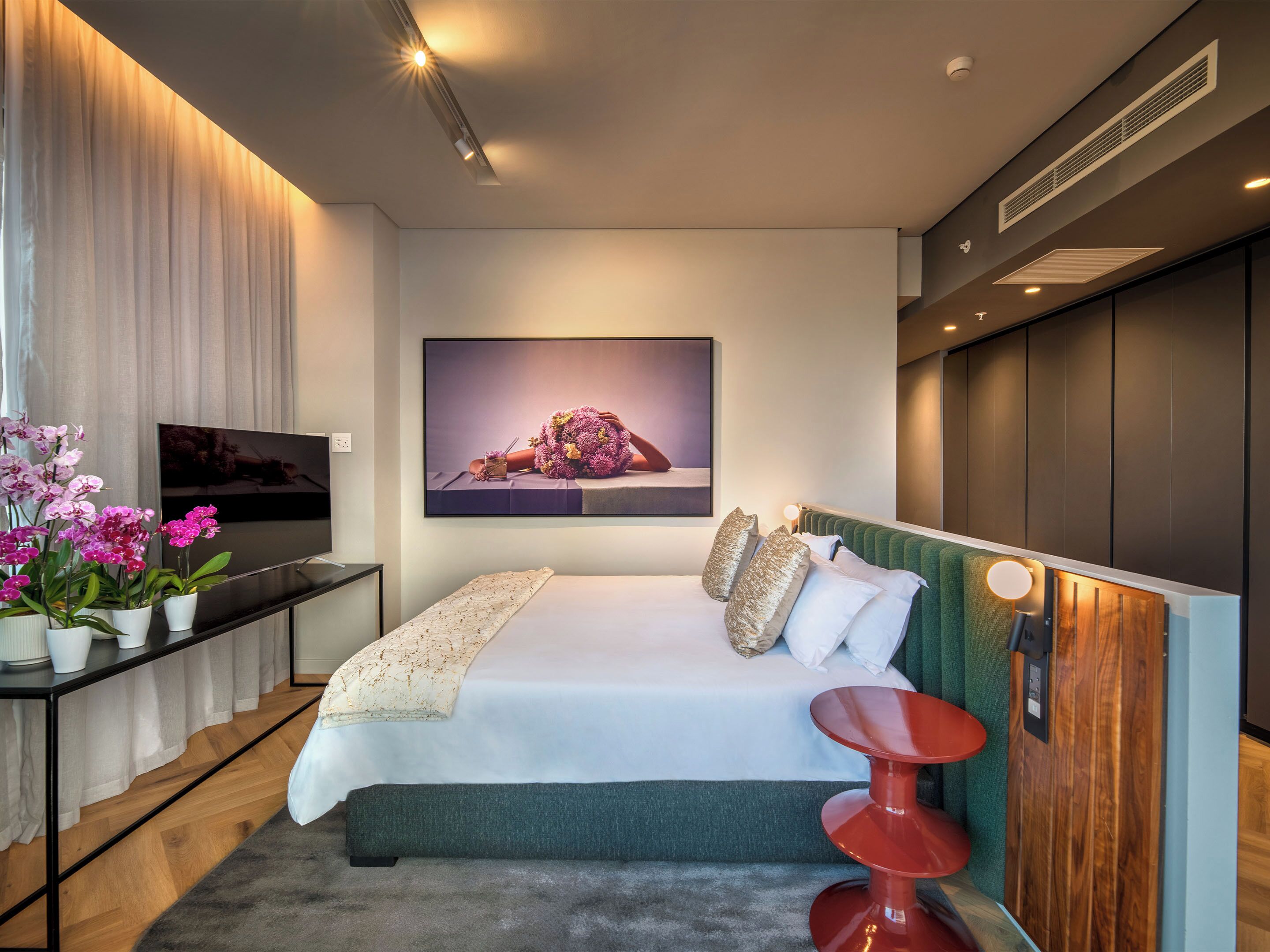 Une chambre moderne avec un lit, une télévision, un tableau et un fauteuil rouge.