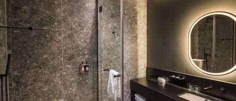 Un baño moderno con ducha envuelta en cristal, un tocador oscuro con espejo redondo y lavamanos integrado.