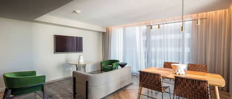 A modern living room with a dining area, a flat-screen TV mounted on the wall, and a large window with sheer curtains.