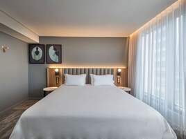 Una habitación de hotel con una cama grande, dos mesitas de noche, un cabecero con iluminación integrada y un ventanal con cortinas translúcidas.