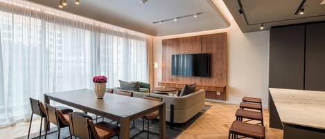 A modern living room with a dining area, a flat-screen TV mounted on a wooden wall, and a kitchen counter.