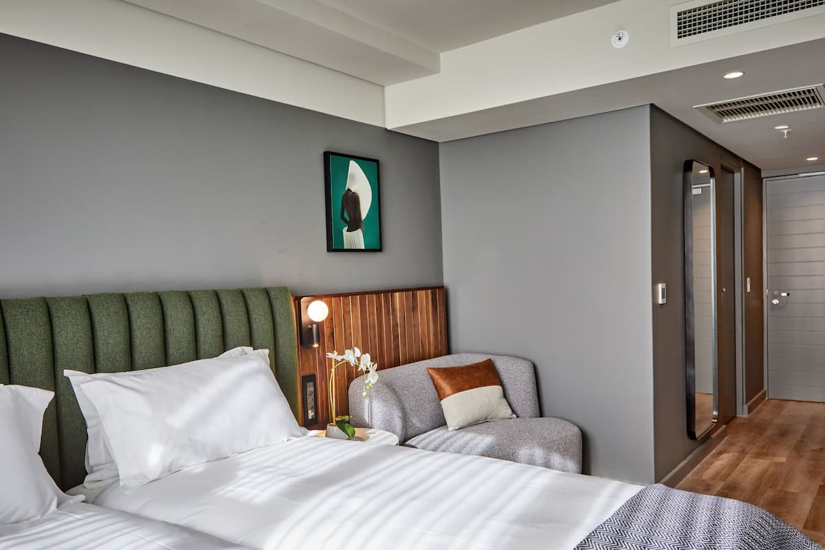 Ein modernes Hotelzimmer mit einem großen Bett, einer Sitzecke mit gepolstertem Sitzmöbel, einem kleinen Tisch mit Blumenvase und einem Gemälde an der Wand.
