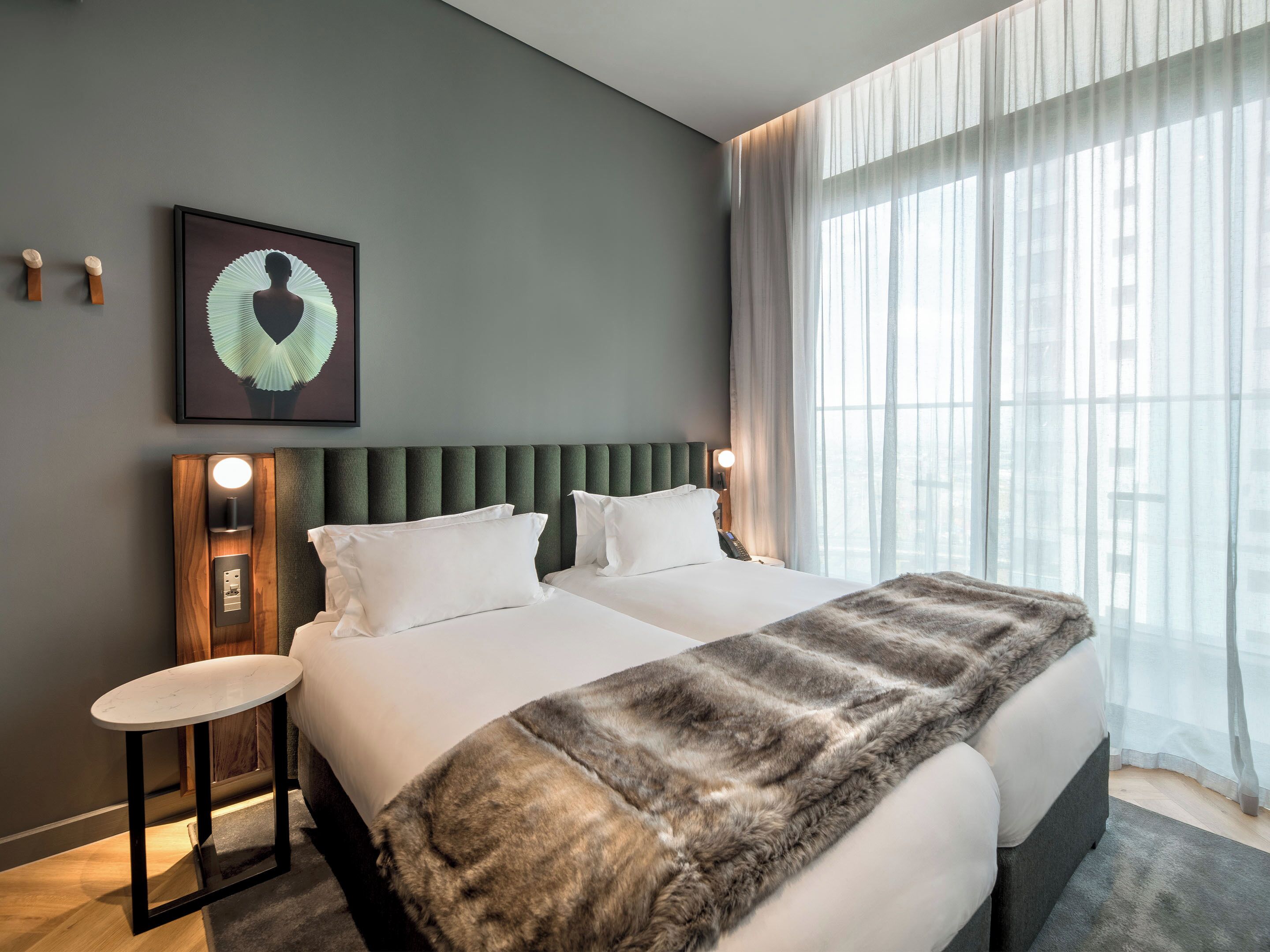 A modern hotel room with a large bed, a small side table, a wall-mounted light, and a large window with sheer curtains.