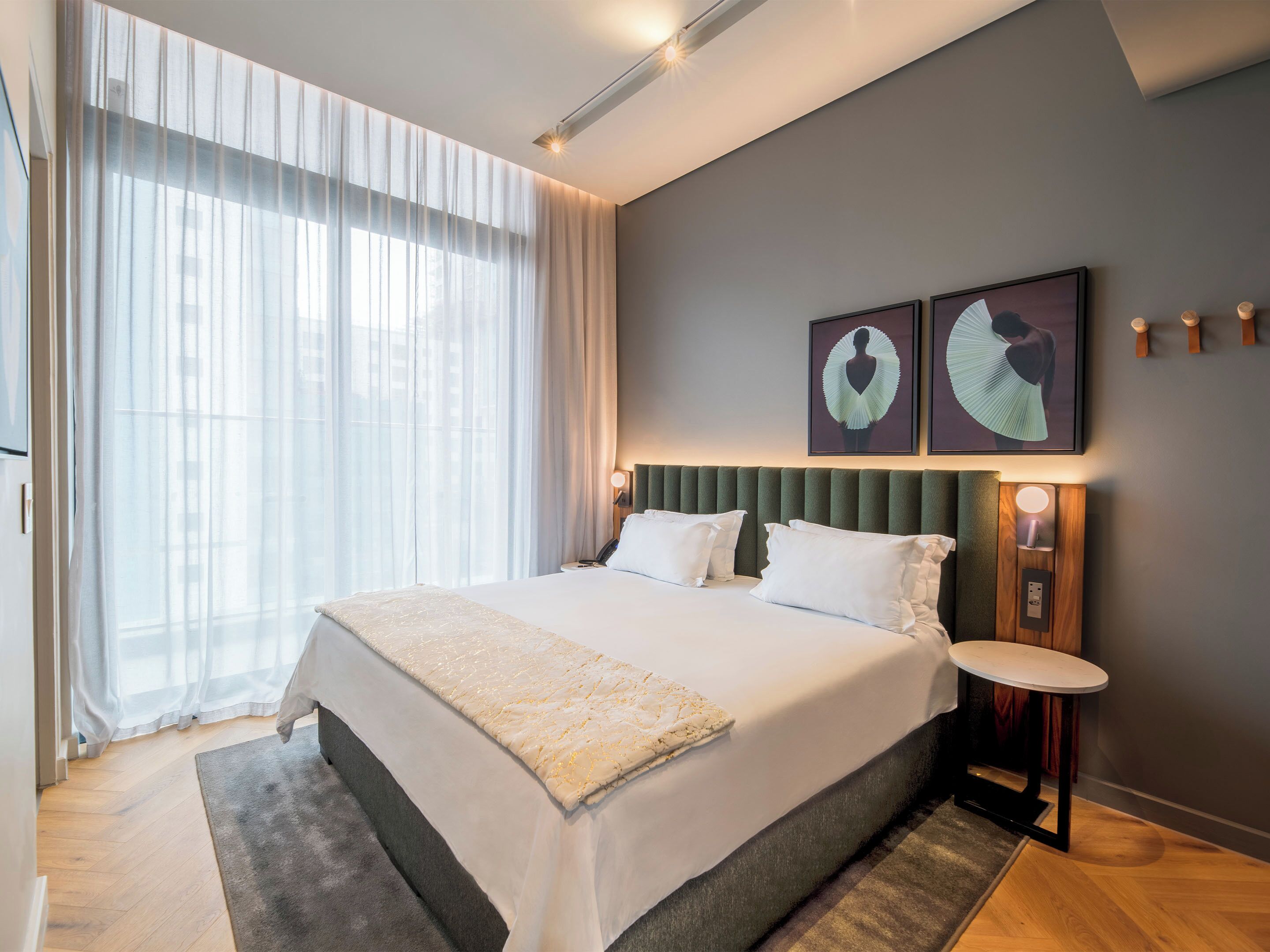 Habitación de hotel moderna con una cama grande, una mesita de noche y vistas de la ciudad a través de cortinas translúcidas.