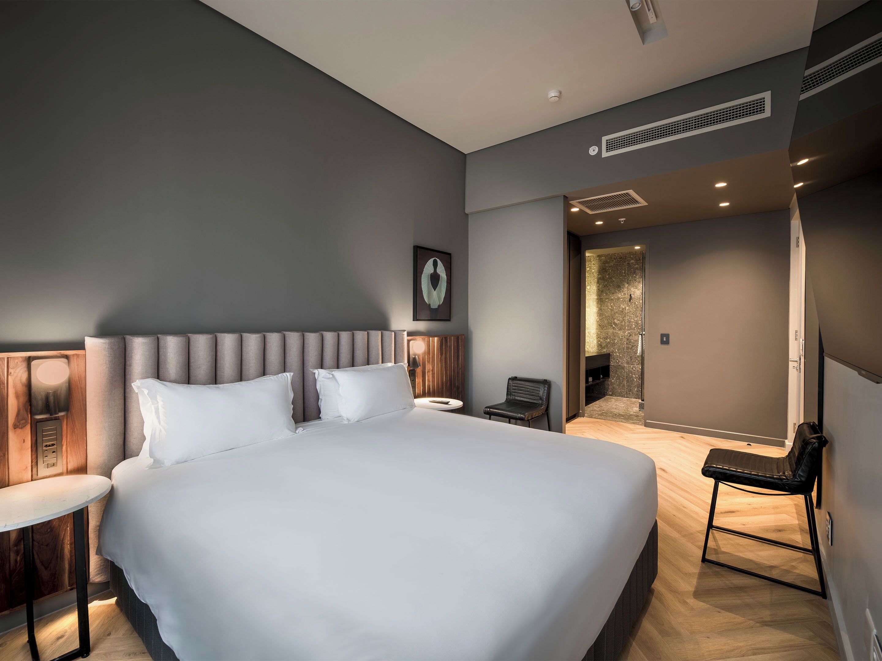 Quarto de hotel moderno com uma cama grande, uma pequena mesa, uma cadeira e um banheiro visível através de uma porta aberta.