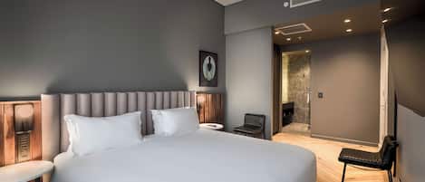 A modern hotel room with a large bed, a small table, a chair, and a bathroom visible through an open door.