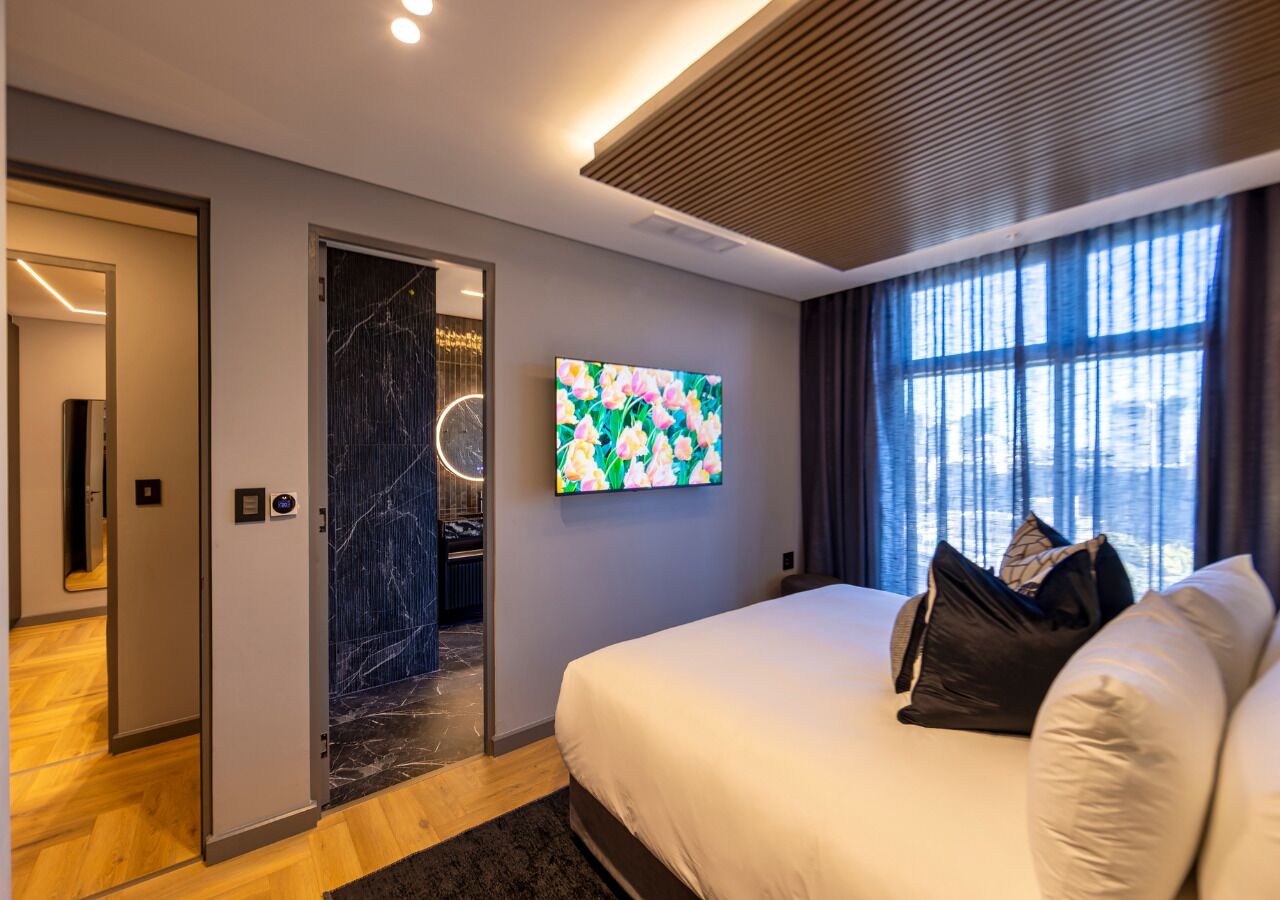 A modern bedroom with a large bed, a flat-screen TV, and a view of the city through large windows.
