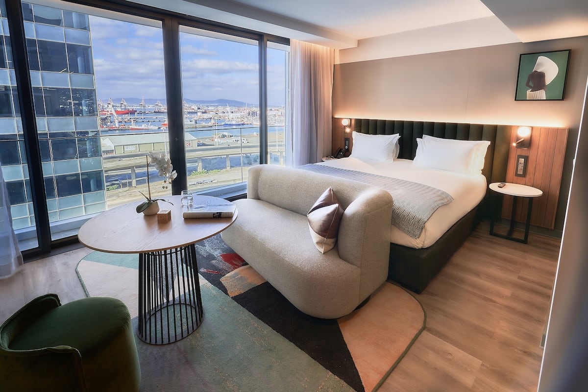 Ein modernes Hotelzimmer mit einem großen Bett, einer Sitzecke, einem kleinen Tisch und Blick auf die Stadt durch große Fenster.