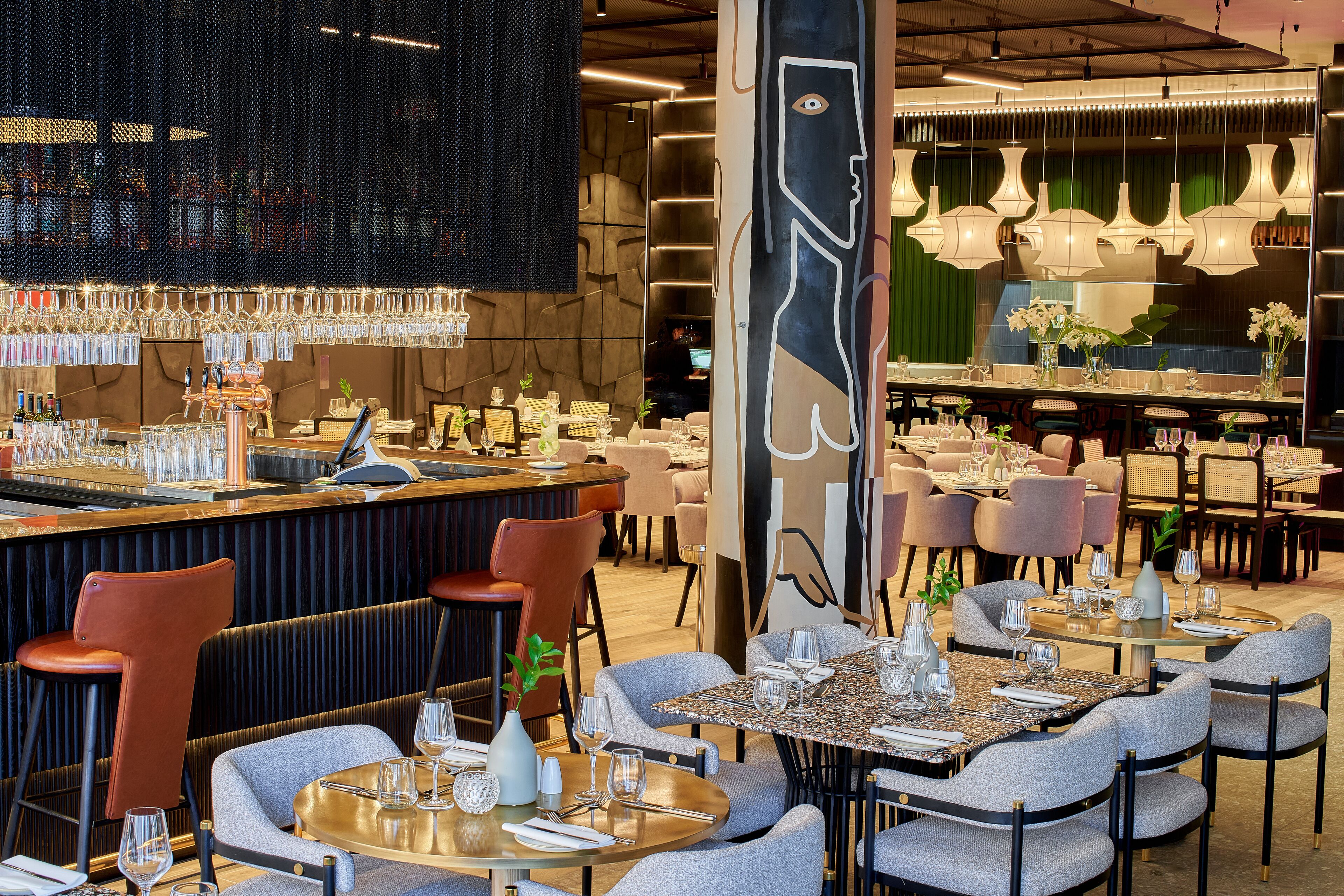 En restaurant med runde borde og barpladser, med et stort muralmaleri og stemningsfuld belysning.