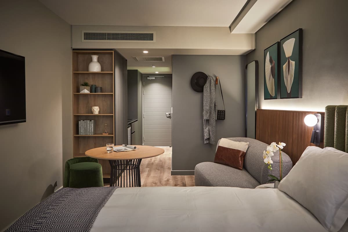Ein modernes Hotelzimmer mit einem Bett, einem kleinen runden Tisch, einem Bücherregal und einem an der Wand befestigten Fernseher.