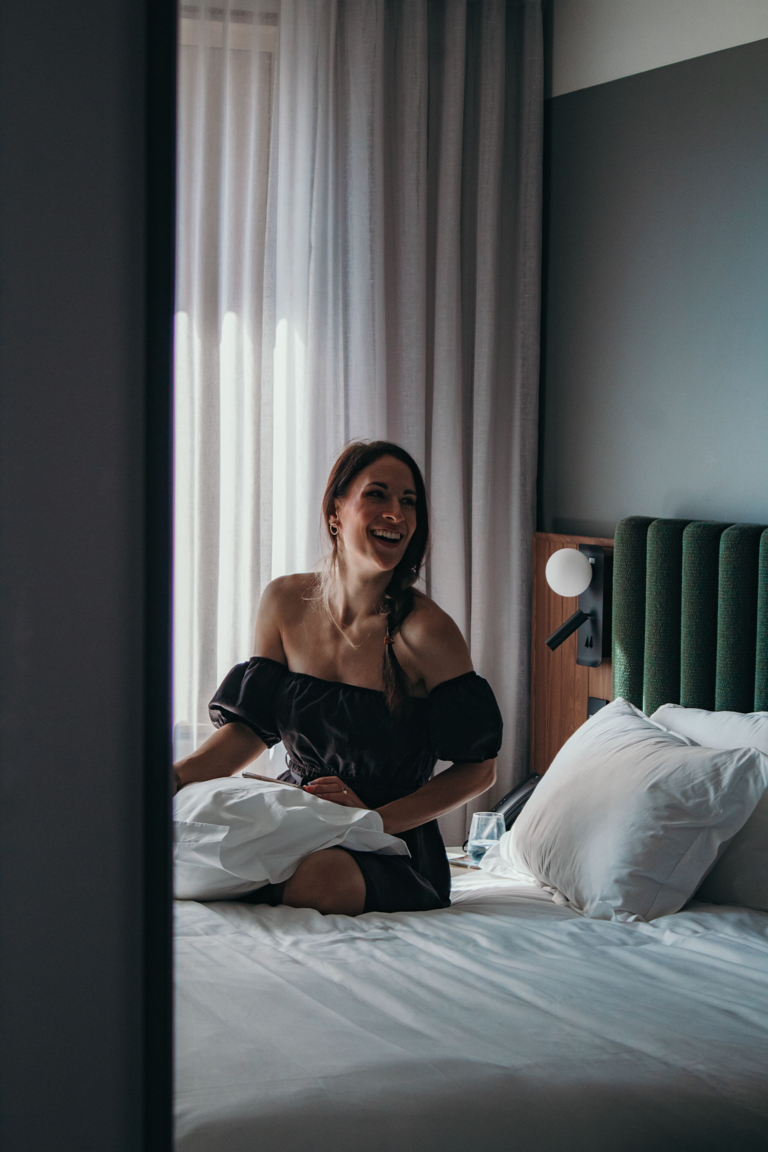 A person sitting on a bed in a room with a green headboard, white bedding, and a window with sheer curtains.