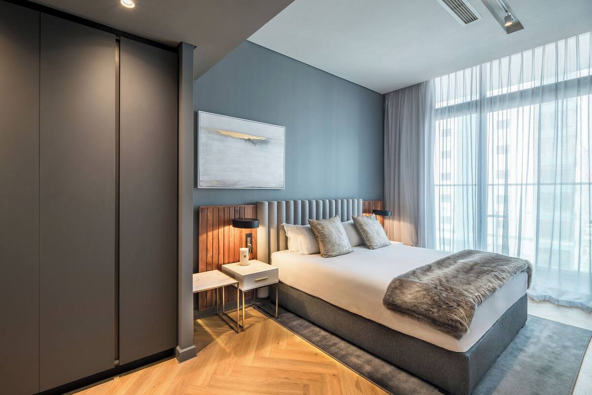 Ein modernes Schlafzimmer mit einem großen Bett, einem Nachttisch, einer Wandlampe und einem Fenster mit transparenten Vorhängen.