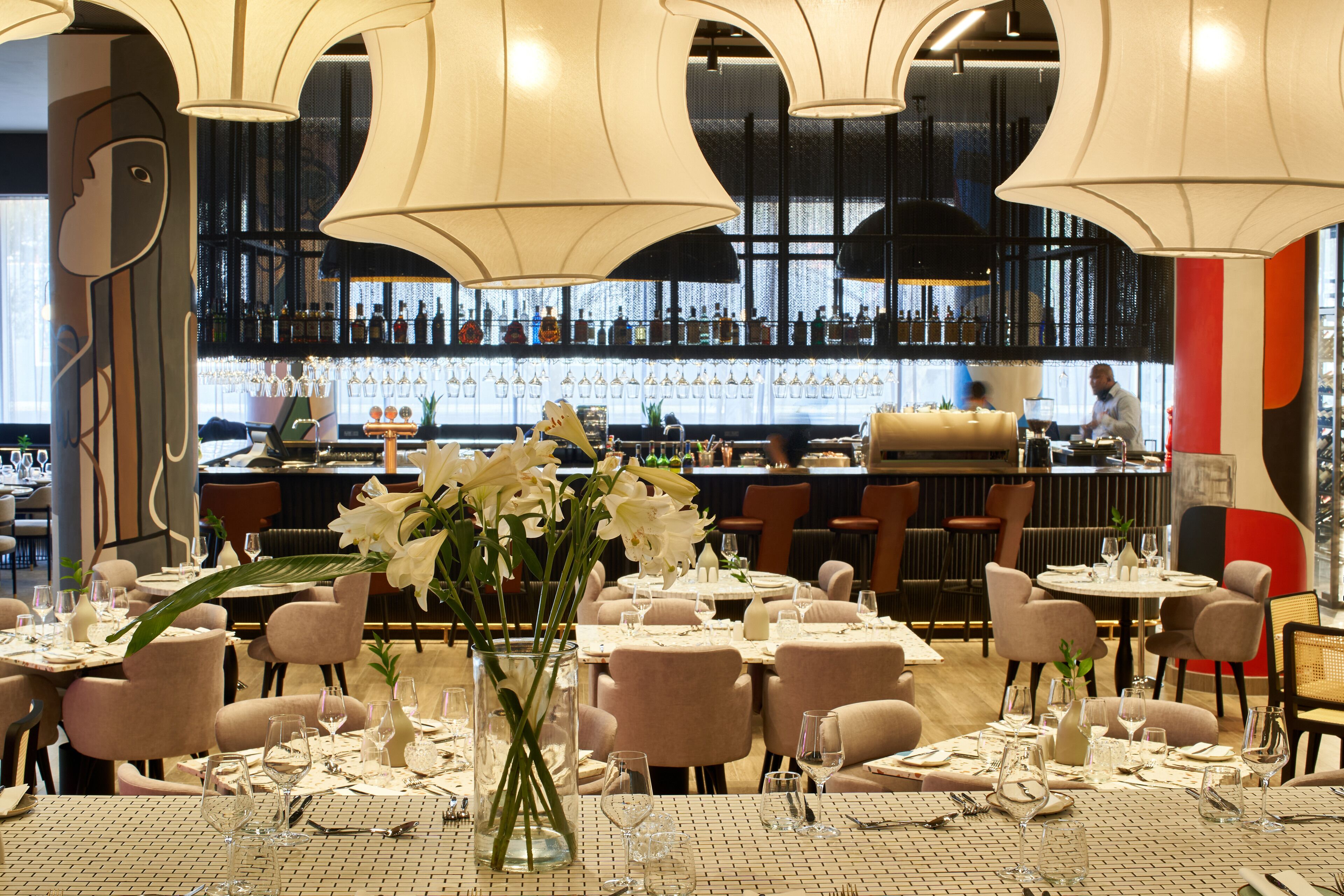 A restaurant with round tables set for dining, a bar in the background, and a large vase of white flowers on the table.