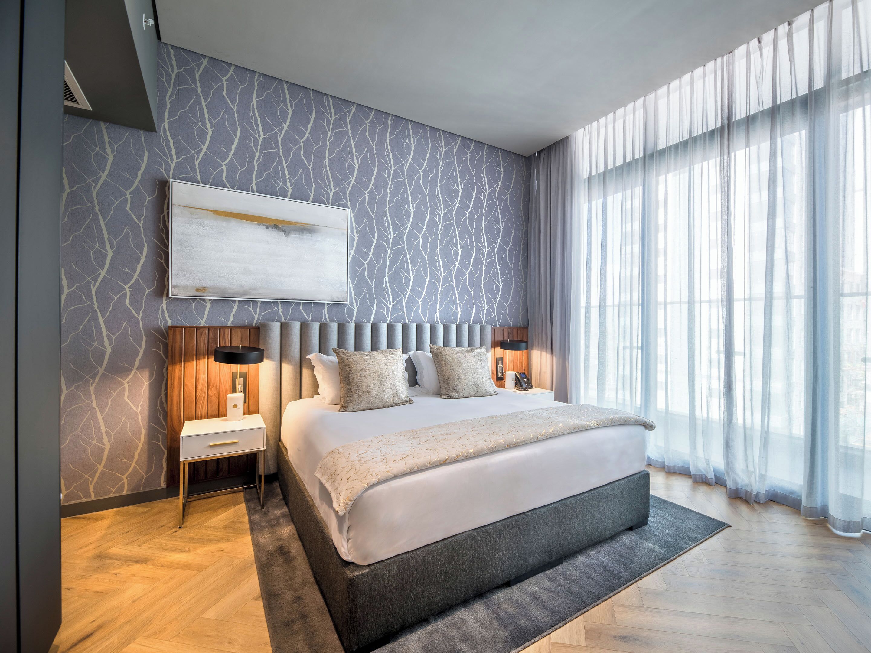 A modern bedroom with a large bed, a nightstand, a wall-mounted lamp, and a large window with sheer curtains.