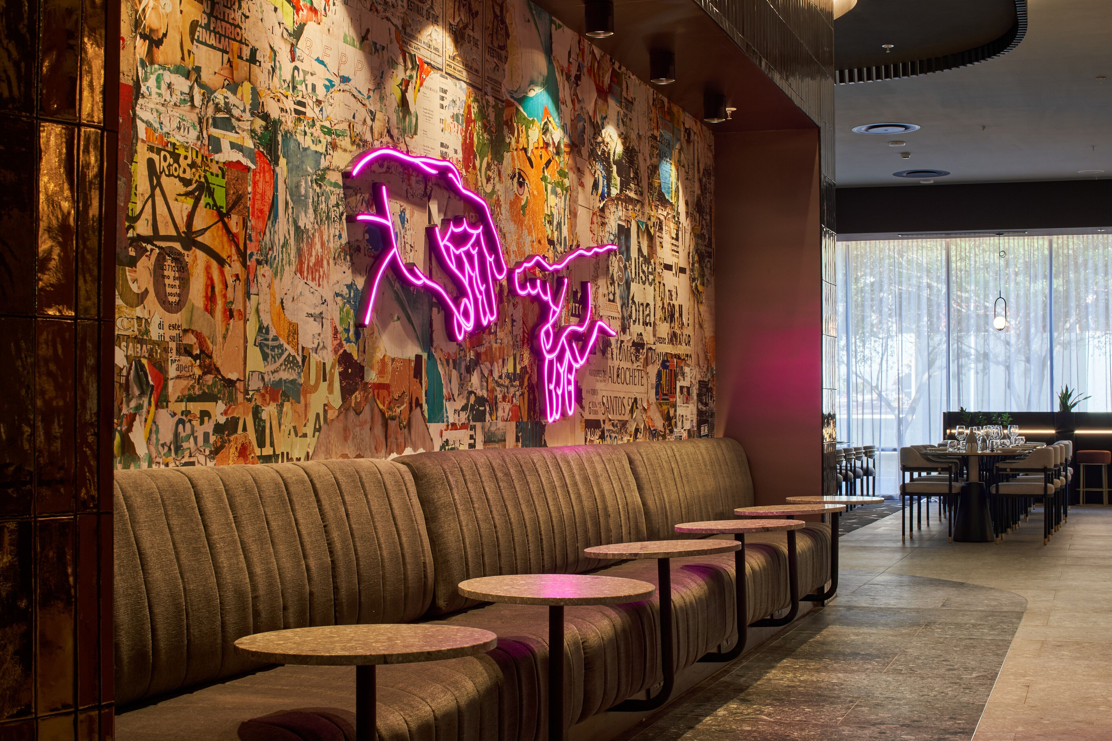 Un restaurante con un letrero de neón, una pared cubierta de carteles y una zona de estar con mesas redondas y bancos acolchados.