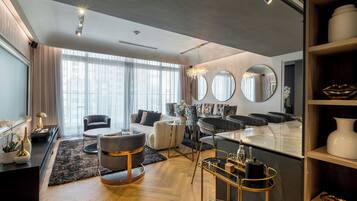Una sala de estar moderna con sofá, sillones, una mesa de centro y una zona de comedor con mesa y sillas. Hay ventanas grandes con cortinas, una lámpara de araña e iluminación empotrada en el techo.